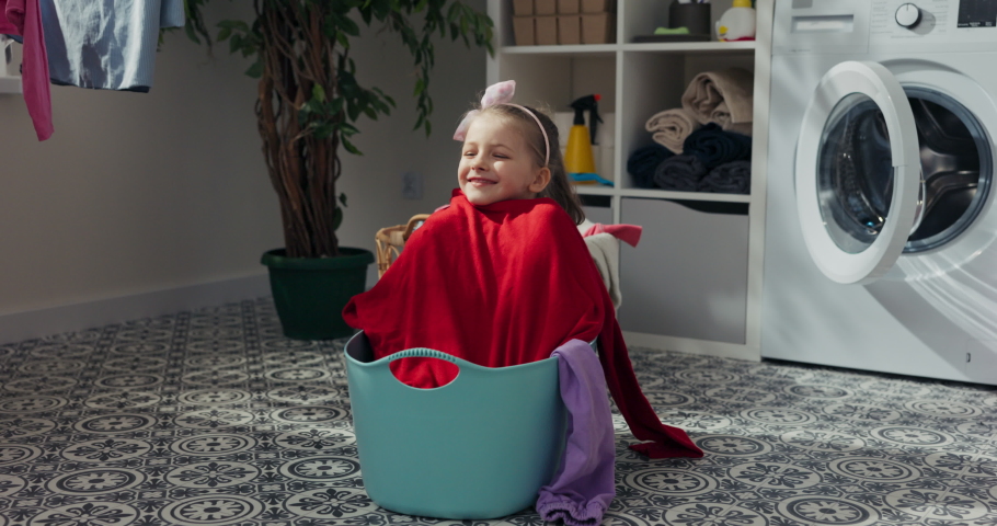 Little girl is sitting in wash basin in bathroom. She pulls out washed clothes and smiles. Girl wrapped herself in freshly washed red sweater. It is soft and smells good. There is washing machine.
