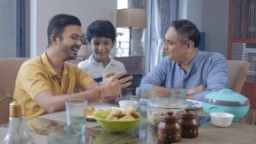 A happy Indian Ethnic young kid boy or grandson, son, and grandfather watching a funny video together on a mobile phone or a smartphone at the dining table before lunch or a meal in an indoor home.