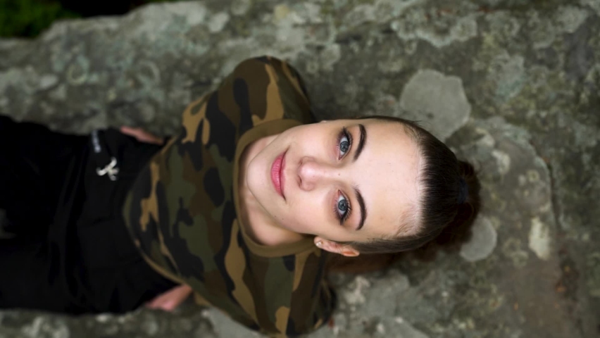 Pretty young girl sits on a stone in the forest, looks at the camera and smiles sweetly. View from above. Vertical video. Social media. 