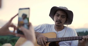 Handheld, Selective focus Young man play guitar and sing a song, pretty woman use smartphone take a photo or record video clip, Young couple have relax time at camping park together
