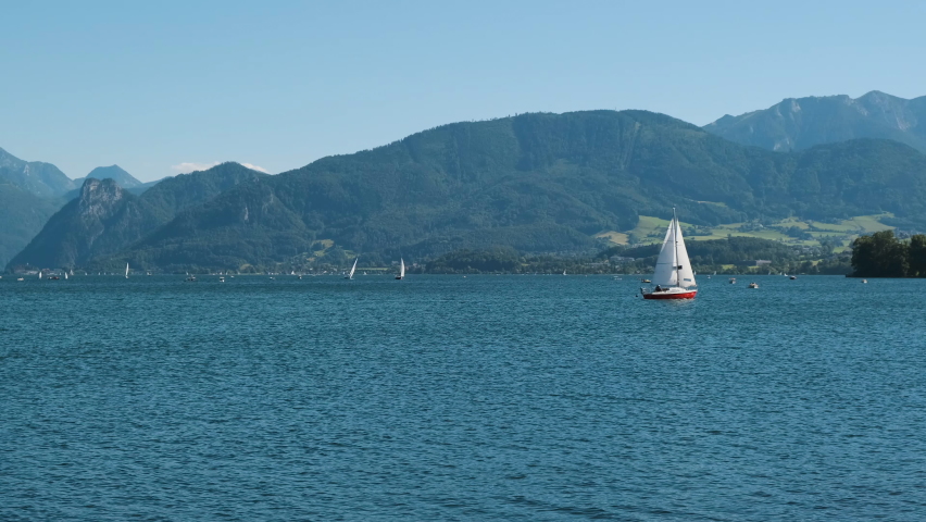 Wide cinematic panorama landscape natural shot with sailing boat on a lake river with clean clear blue water on gorgeous green forest mountains background. Static camera view of a summer vacation day