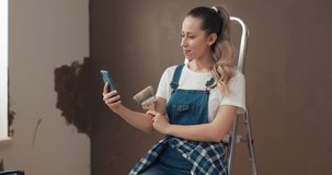 Woman in denim overalls is sitting on ladder, holding phone and brush in hands and calling friend. She has video call and shows new place. Girl with curly hair is talking and smiling.