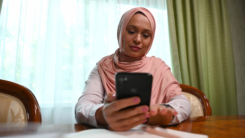 Charming Middle-Eastern Muslim woman in pink hijab, using smartphone while relaxing at home, feeling shocked, stressed and disappointed, reading bad news on her mobile phone, feeling fear and sadness | Shutterstock HD Video #1095407815