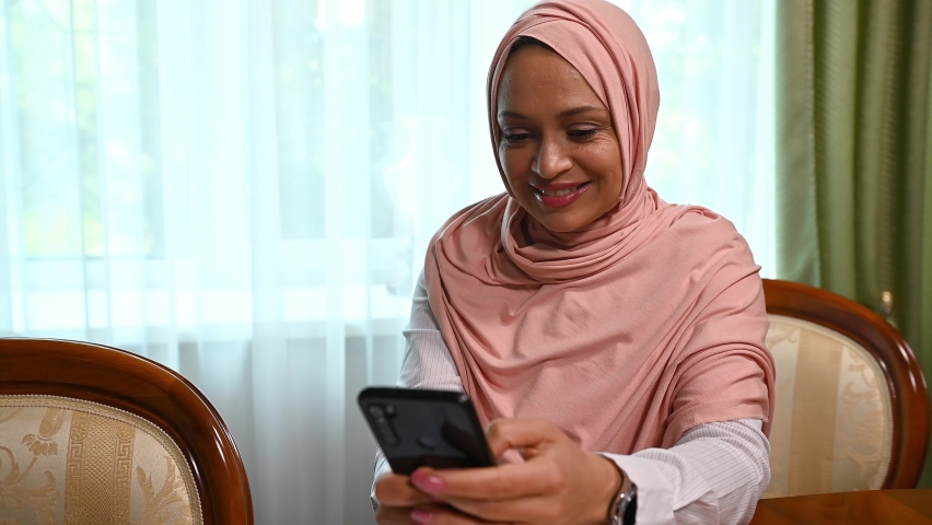 Charming Arab Muslim woman wearing pink hijab, relaxing in living room, using smartphone, checking content, looking away dreamily, smiling a beautiful toothy smile, working remotely, texting messages | Shutterstock HD Video #1095407821