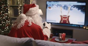 Happy Santa Claus sitting on the sofa at home and playing video games at Christmas