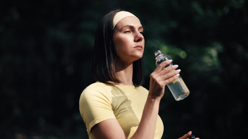 Active young adult woman drinking water from glass bottle. The fitness female take a rest outdoor. Concept of workouts running and healthy lifestyle. Yellow and green. Royalty-Free Stock Footage #1095412063