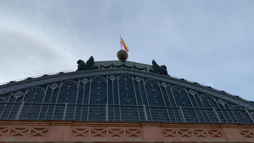 Spanish flag is waiving on top of Madrid Atocha train, railway station building roof. Main, front entrance. Bright blue sky. Madrid, Spain
