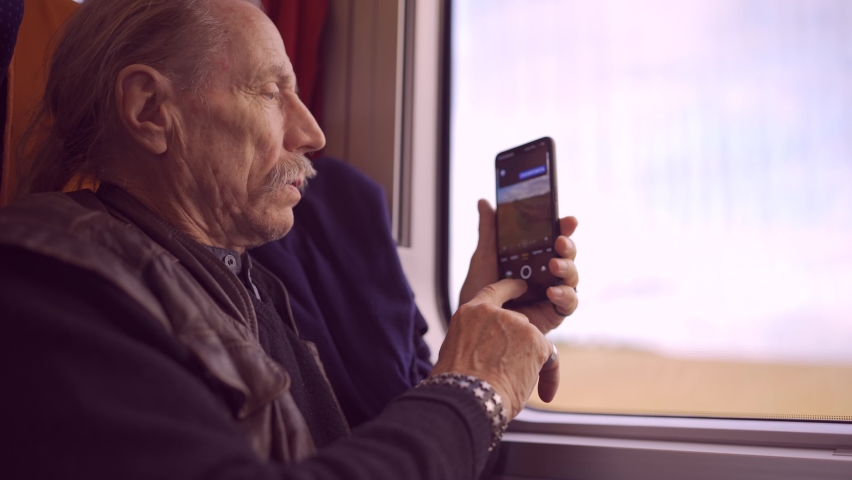 Elderly man traveling by train and taking pictures of the landscape through the window using a smartphone