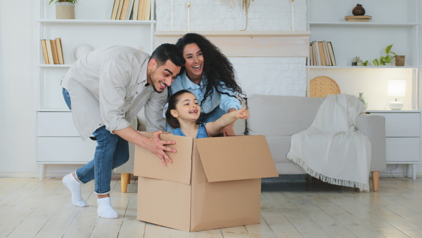 Multiracial multiethnic mother and father Indian Hispanic Caucasian parents push cardboard boxes with little cute child girl daughter ride inside homeowners family playing fun in new house relocation