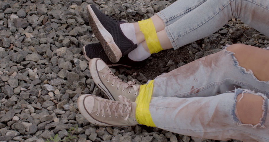 Kidnapping. Two unrecognizable women lie helplessly on the ground, experiencing fear, dirty torn jeans. Close-up of bound female legs with yellow tape. Escape plan. Get into no-win situation. Outdoors