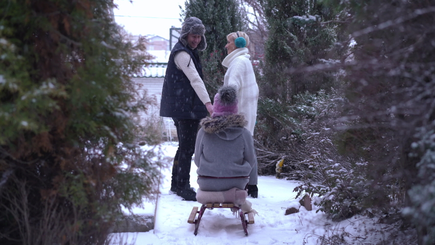 Back view wide shot of joyful teen girl stretching hands as man and woman pulling sled in slow motion smiling. Happy carefree Caucasian daughter having fun with parents on winter backyard outdoors