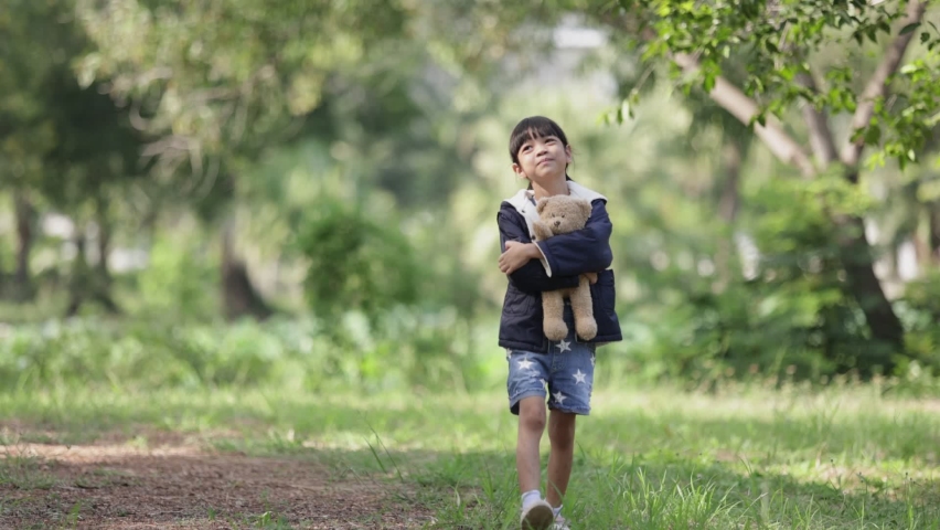 Portrait, a beautiful Thai Asian kid girl aged 5 to 7,wearing a blue sweater and holding a teddy bear.Take a walk in the outdoor garden happily The background is green trees. Autumn diversity concept.
