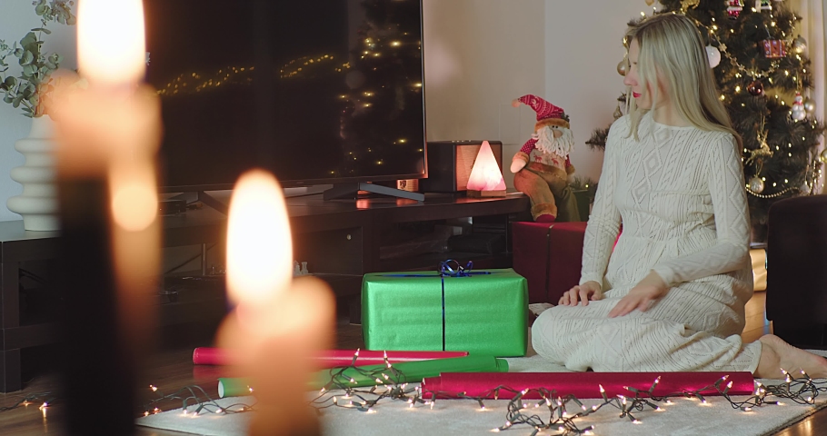 Woman wrapping gifts at Home Christmas Eve in Winter Day, Mother wrapping a gift in gift paper while sitting on the floor in the room. Holiday concept, color full lights bokeh background.