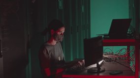 High angle of female Caucasian programmer wearing face mask sitting at desk in dark green-lighted server room, typing on computer - Powered by Shutterstock - Get 15% off with code: PIKWIZARD15