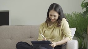 happy asian woman relaxing at home sitting on sofa holding digital tablet enjoying internet surfing study work online shopping