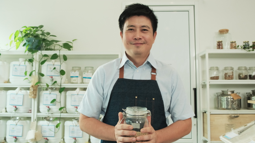 Portrait of Thai male shopkeeper smiles and looks at camera with glass jar of natural organic products at refill store, zero-waste grocery, and plastic-free, environment-friendly sustainable business.