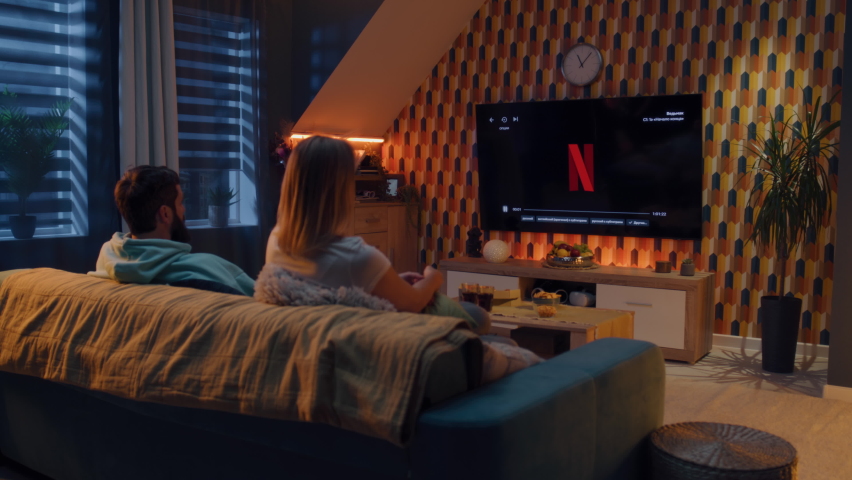 ZAPORIZHYA, UKRAINE - October 13, 2022: Couple sitting on sofa together, watching movie or TV series on Netflix streaming service with snacks, soda, and pizza. Man and woman chilling resting at home.