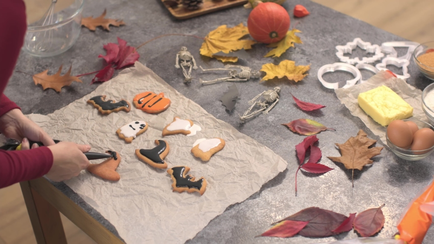 decorating halloween black witch hat cookies with colorful glaze royal icing autumn orange pumpkins yellow leaves on kitchen table. black bat white ghost shape biscuits celebrating halloween party