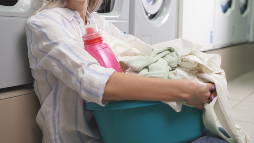 The washing machine of the public laundry washes dirty colored clothes, and an attractive woman sits by the car waiting for clean laundry.