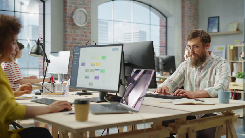 Portrait of Creative Bearded White Man with Glasses Smiling While Using Computer in Bright Spacious Office. Male Team Manager Giving Feedback to colleagues. People Working in Background. Dolly Shot.