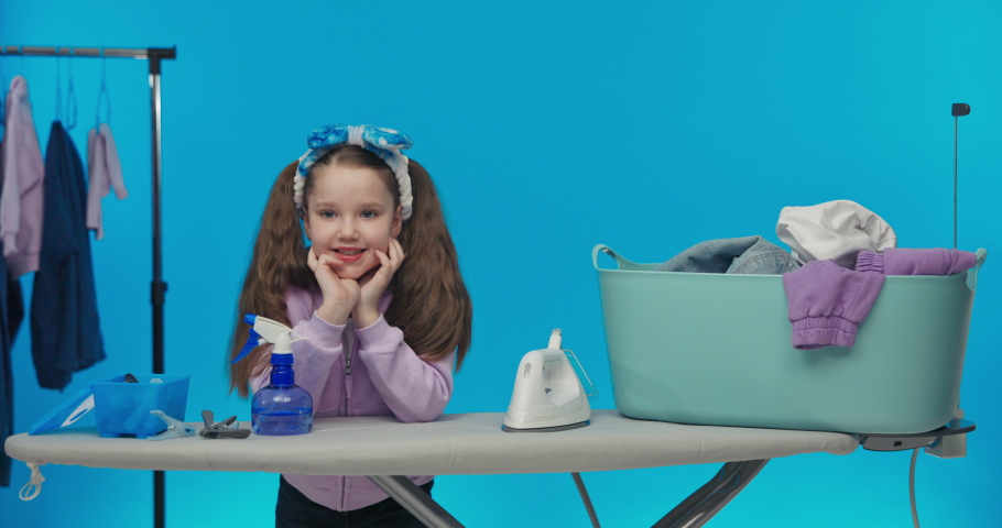 Laundry room on a colorful blue studio background. Small smiling sweet girl is doing household chores, sorting laundry, preparing for ironing, holding phone in hand.
