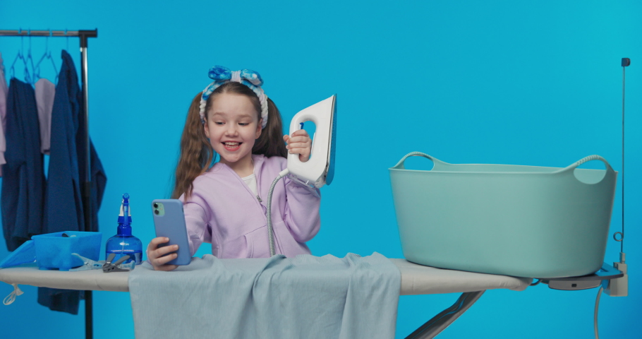 Happy smiling little girl little housewife ironing clothes laundry iron at home on blue background studio.