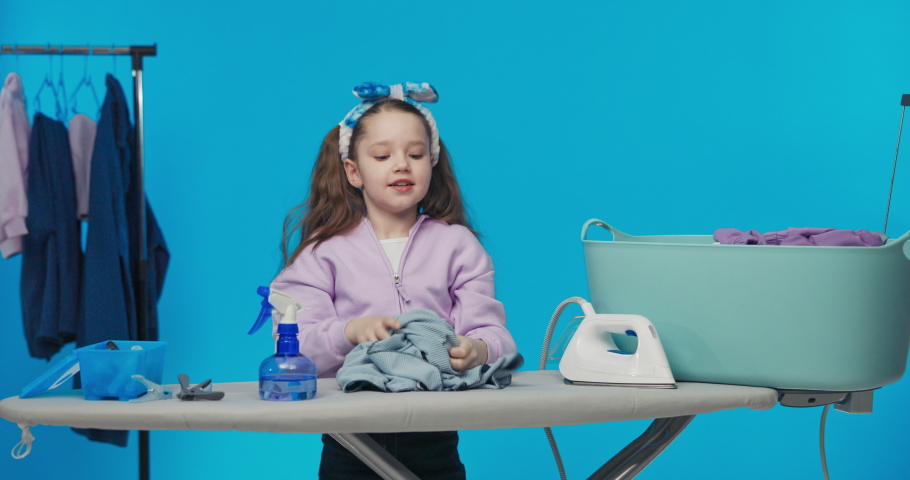 Little cute girl in headband and two ponies stands on blue pastel studio background folds clothes on board takes them out of big bowl, preparation for ironing.