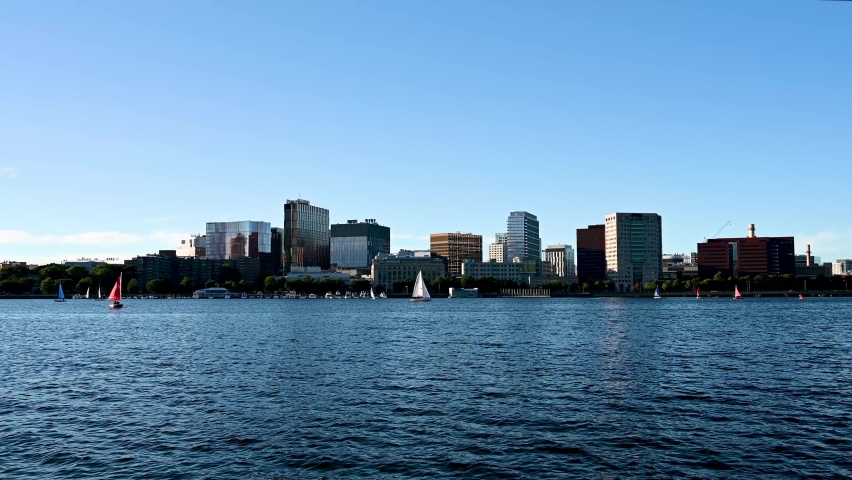 The Boston skyline under a blue sky. Sailboats sail across the Charles River.