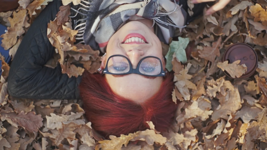a girl with red hair lies on the fallen leaves of trees and smiles. slow motion video