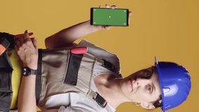 Vertical video: Powerful woman contractor pointing at greenscreen display on smartphone, standing over yellow background. Using isolated copyspace with mockup template and chroma key, building