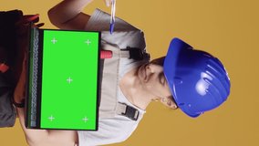 Vertical video: Portrait of handywoman holding laptop with greenscreen display over yellow background, using blank chroma key template with isolated mockup copyspace. Working on renovation with