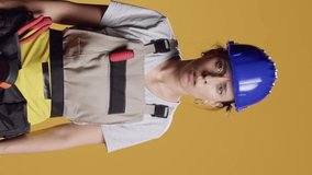 Vertical video: Portrait of confident handywoman acting curious and exploring, standing over yellow background. Female contractor looking at camera and being silly, investigating and searching