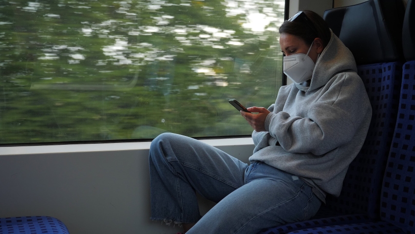 Young woman traveling by train, holding smartphone in hands, reading electronic book. Female traveller sitting in train commuting to work. Woman in protective mask rides public transport