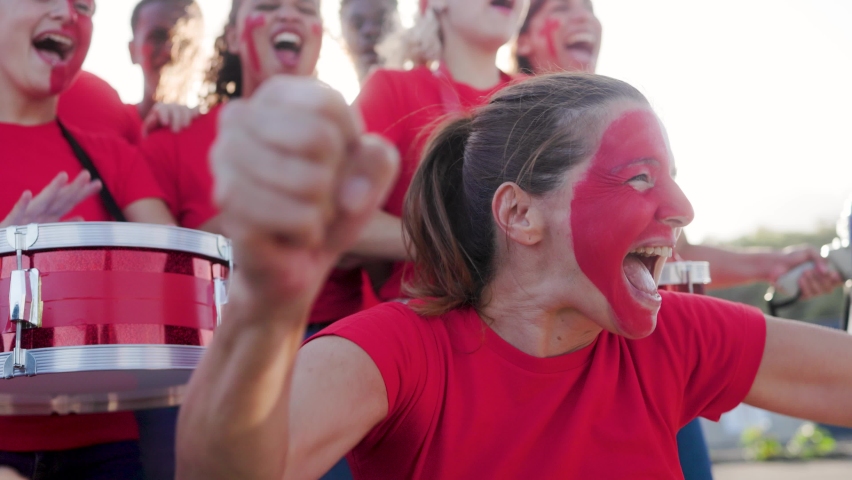 Multiracial football fans celebrating and supporting their red sport team watching the game on stadium tribune - Soccer supporters screaming during event.