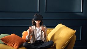 A teenage girl in virtual reality glasses or 3d glasses sits on a yellow sofa at home. People entertainment concept. Technology, virtual reality, cyberspace.