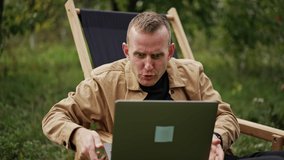 Emotional talk through the video chat on laptop. Man holding computer on his laps sitting in a garden chair outdoors. Apple garden blurred backdrop.