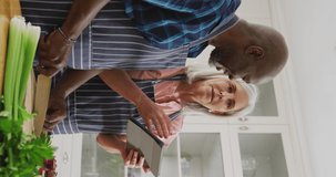 Vertical video of happy diverse senior couple preparing food together using tablet in kitchen. Communication, happiness, health, retirement, domestic life and inclusivity concept.
