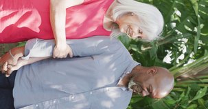 Vertical video of happy diverse senior couple walking in garden arm in arm talking. Happiness, health, retirement, togetherness, love, domestic life and inclusivity concept.