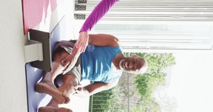 Vertical video of happy diverse senior couple practicing yoga exercising together sitting indoors. Happiness, health, retirement, togetherness, domestic life and inclusivity concept.