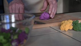 Side view of person cutting purple cauliflower on wooden cutting board on table in domestic kitchen. Real time video. Selective focus. Home cooking theme.