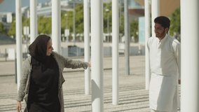Young muslim couple playing hide-and-seek game between columns on sunny day. Arabic man in traditional clothes and woman in hijab having fun, dancing and flirting outside. Romance, ethnicity concept. 