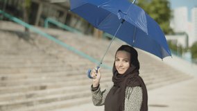 Young Muslim woman in hijab holding umbrella over her head. Smiling Arabic girl lifting up opened blue umbrella, then spinning it, looking at camera. Medium shot. Fashion, lifestyle concept. 