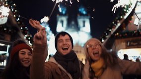 Three happy friends with sparklers on new year's eve to the sound of chimes in the middle of the street in the lights of the Christmas market against the background of the Christmas tree. Film Shot