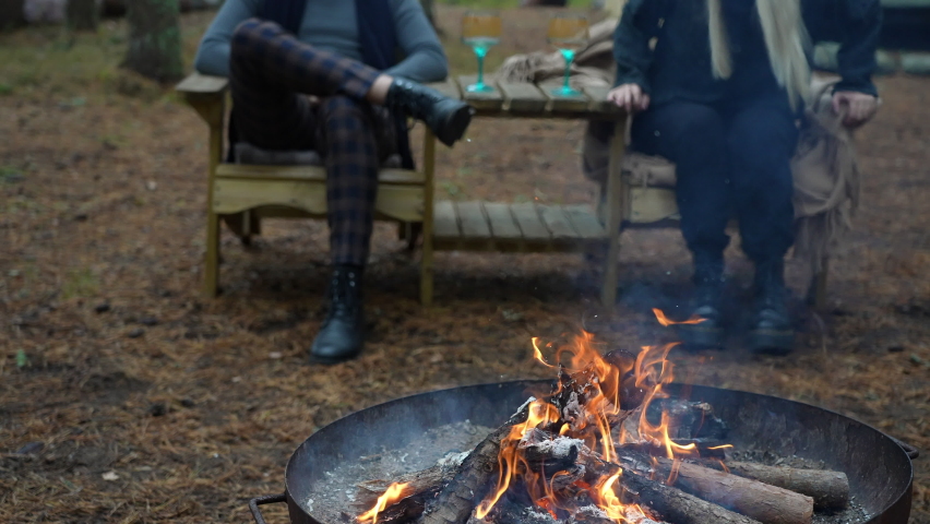 Scenic outdoor bonfire burning flames during a cold beautiful autumn day in the Estonian forest by a wooden cabin with a happy couple sitting in chairs in the background cozy romantic drinking wine Royalty-Free Stock Footage #1095523821