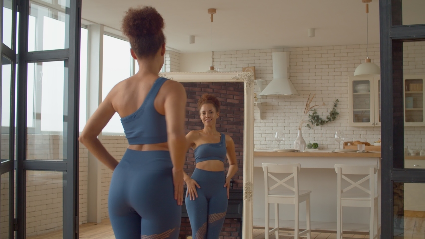Reflection in mirror of slim smiling attractive African woman in sportswear looking at body shape, rejoicing success of dieting plan and weight loss achievement, expressing cheerful mood and happiness Royalty-Free Stock Footage #1095526009