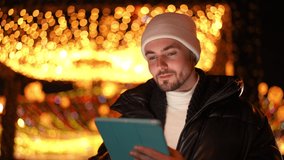 Young Caucasian positive man scrolling social media in digital tablet application looking around at night outdoors. Portrait of absorbed millennial guy messaging online on Christmas eve in urban city - Powered by Shutterstock - Get 15% off with code: PIKWIZARD15