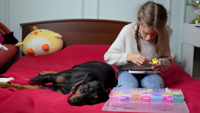 Kid girl watches educational videos on phone and weaves accessories from rubber bands with slingshot and hook at home. Girl with dog lying next to her and accessories for weaving is sitting on bed