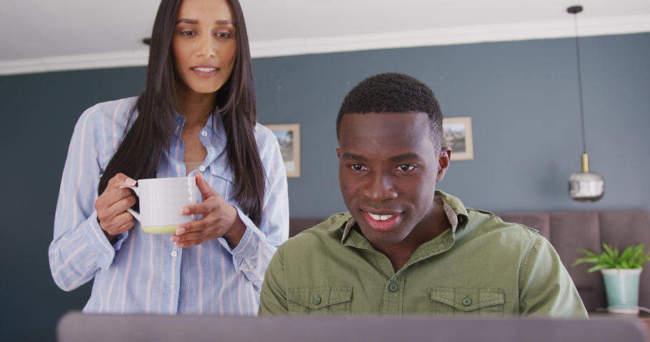 Video of happy diverse couple in bedroom drinking coffee and looking at laptop screen together. Communication, domestic life, and inclusivity concept.