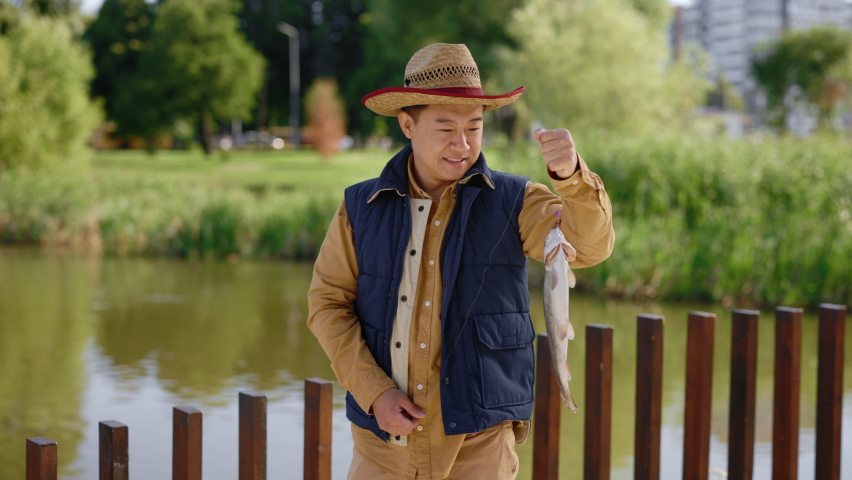 Asian man wearing special fishing clothes holding the fish he caught on the hook and feeling happy about it looking at the camera