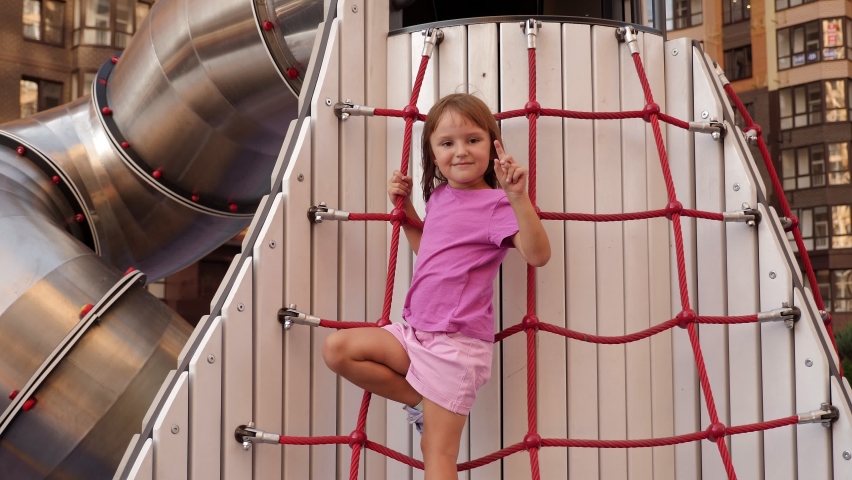 A little girl is playing on a modern playground in the courtyard of a new residential complex, she is climbing a rope ladder.